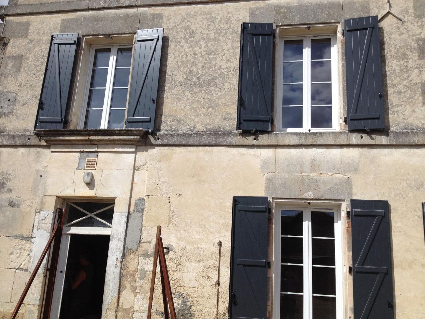 Bâtiment en pierre avec volets noirs sur des fenêtres à cadre blanc ; porte ouverte avec poutres de soutien.