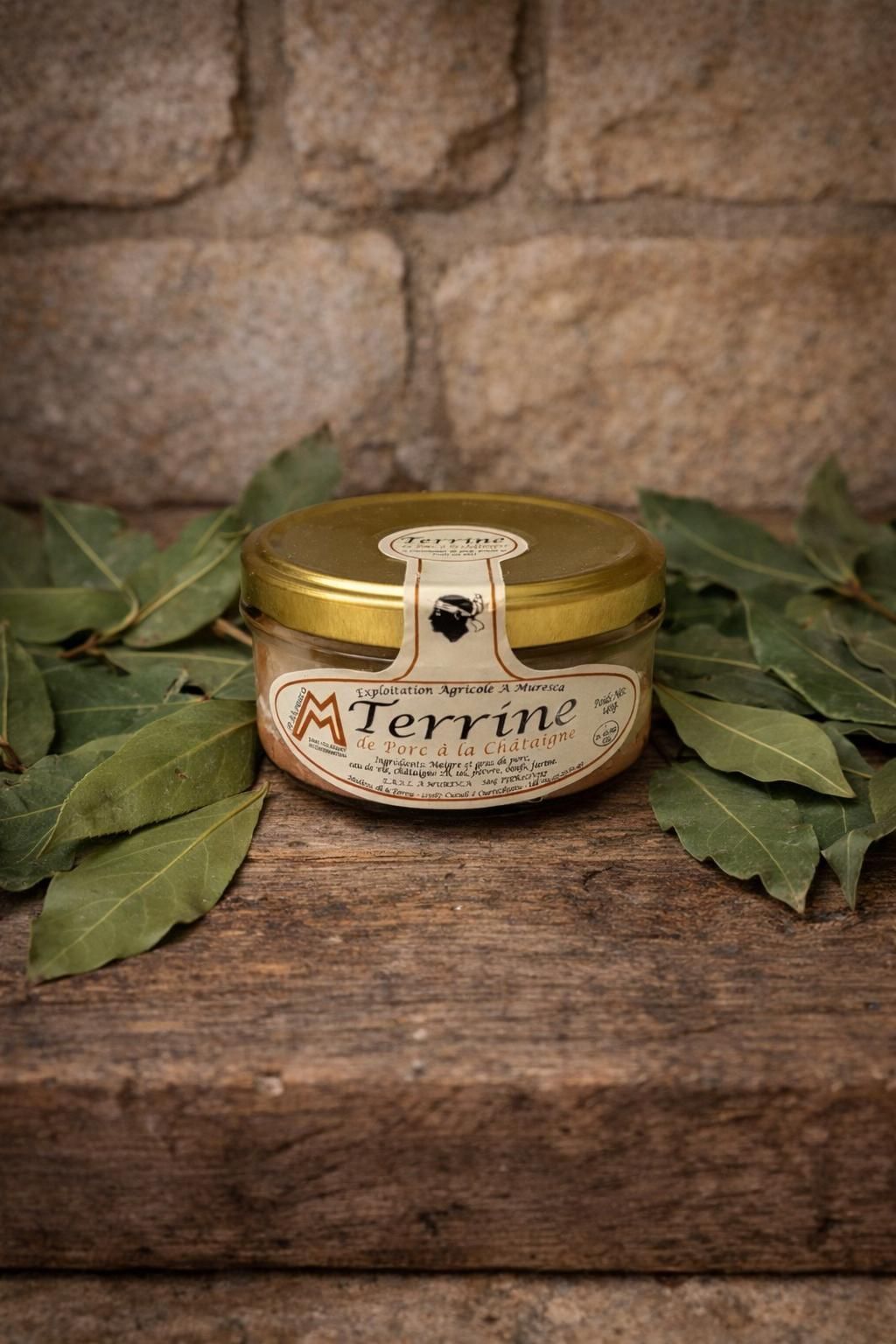 Un bocal en verre contenant une terrine, coiffé d'un couvercle doré, repose sur une surface en bois, entouré de feuilles de laurier sur un fond de pierre.