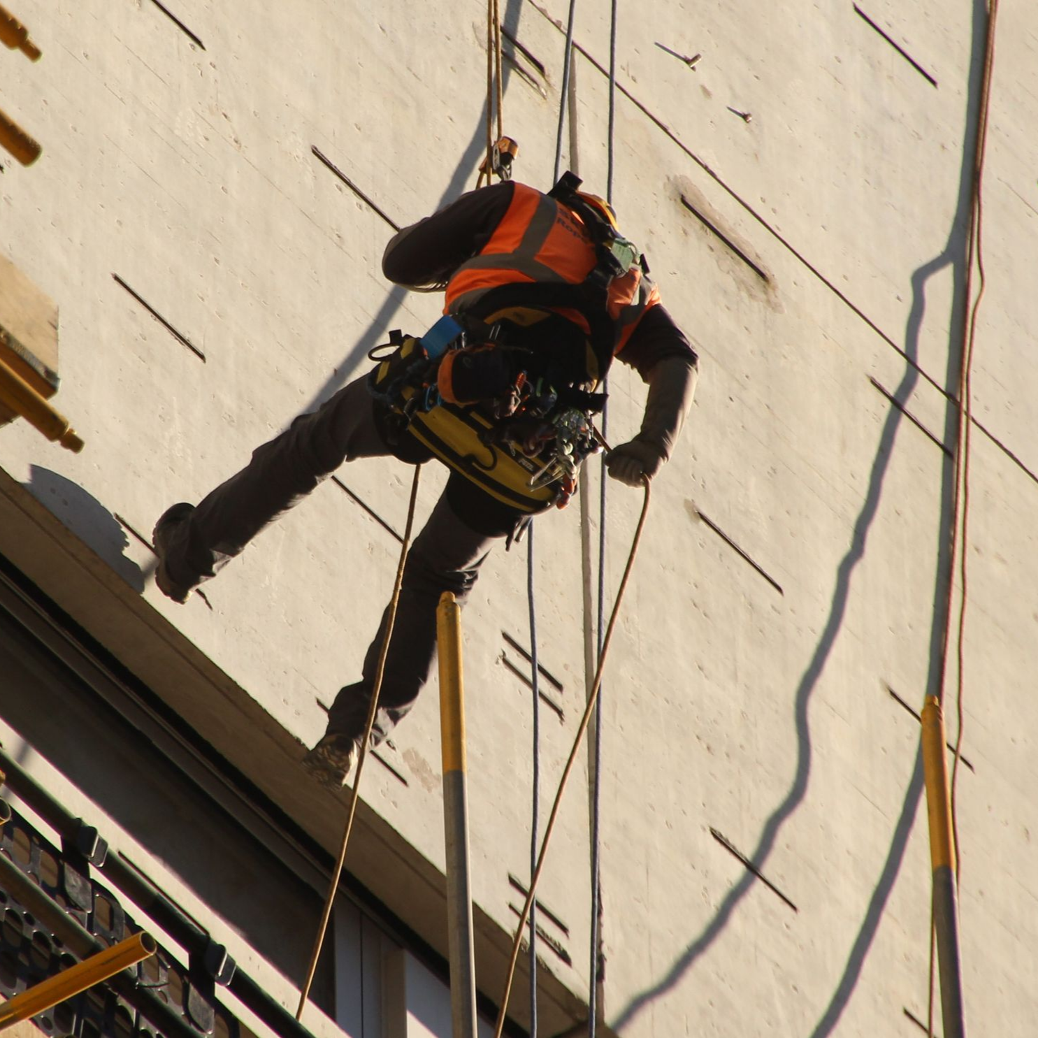 Un ouvrier du bâtiment portant un gilet orange descend en rappel le long d'un bâtiment blanc.