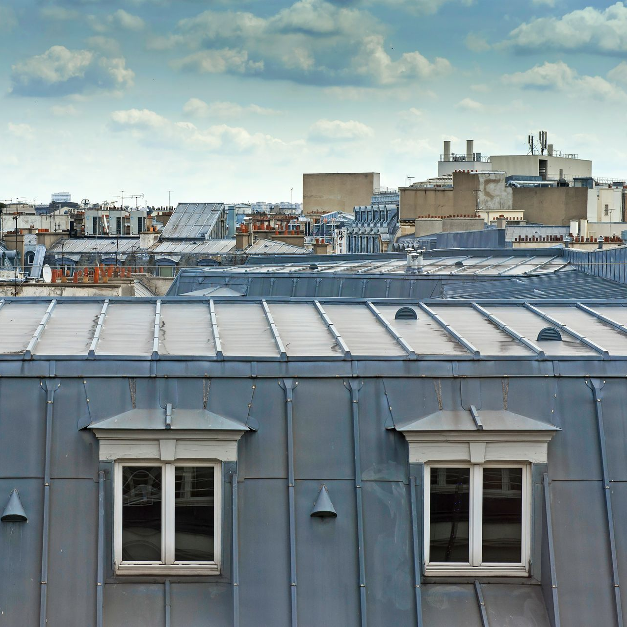 Vue des toits des immeubles parisiens sous un ciel nuageux ; un toit en métal gris avec deux lucarnes est au premier plan.