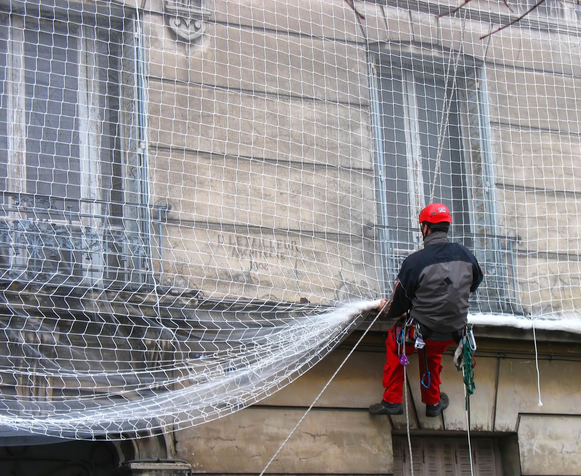 Homme en pantalon rouge et casque sur des cordes, travaillant sur un filet sur la façade d'un bâtiment.