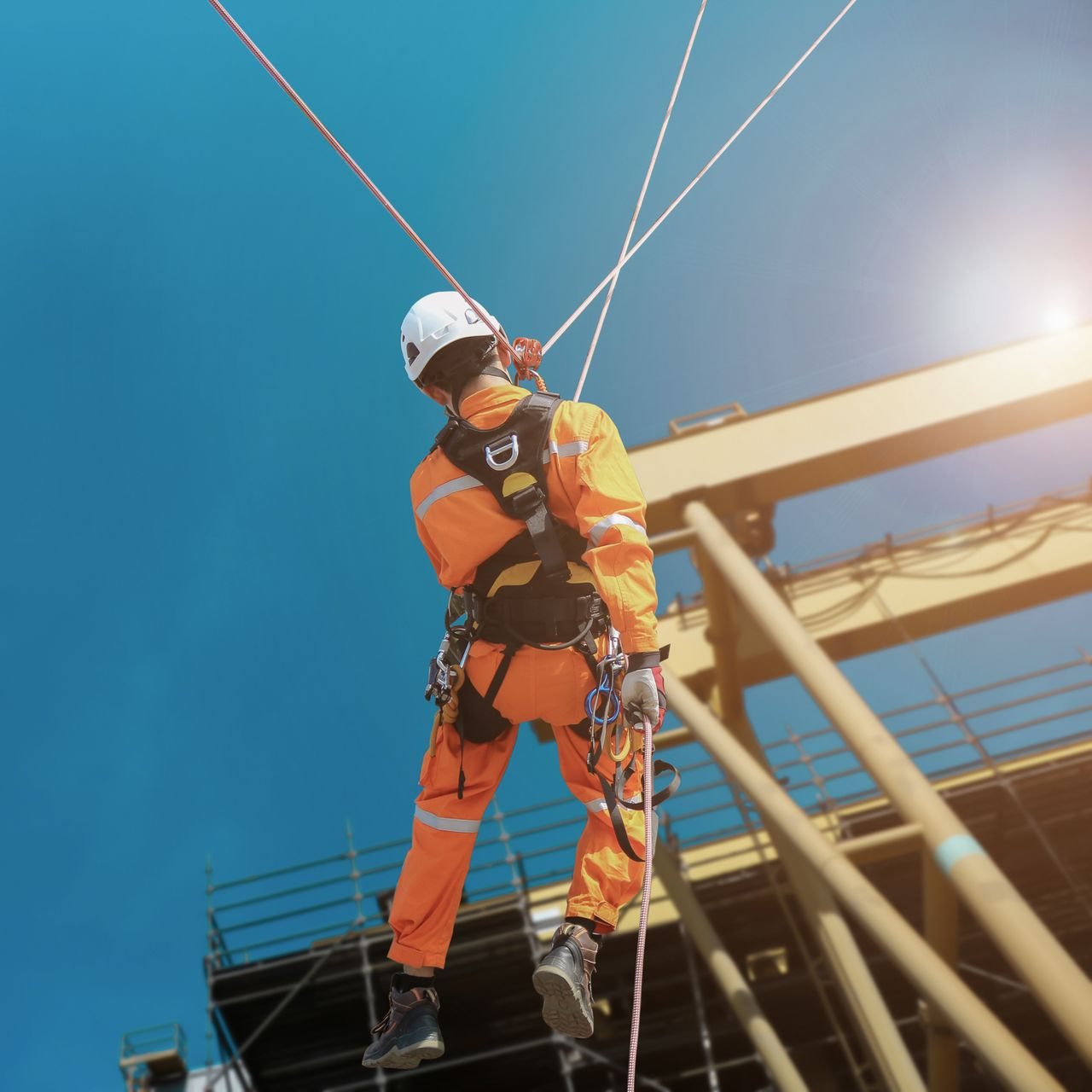 Un ouvrier en tenue de sécurité orange descend en rappel d'une haute structure sous un ciel bleu.
