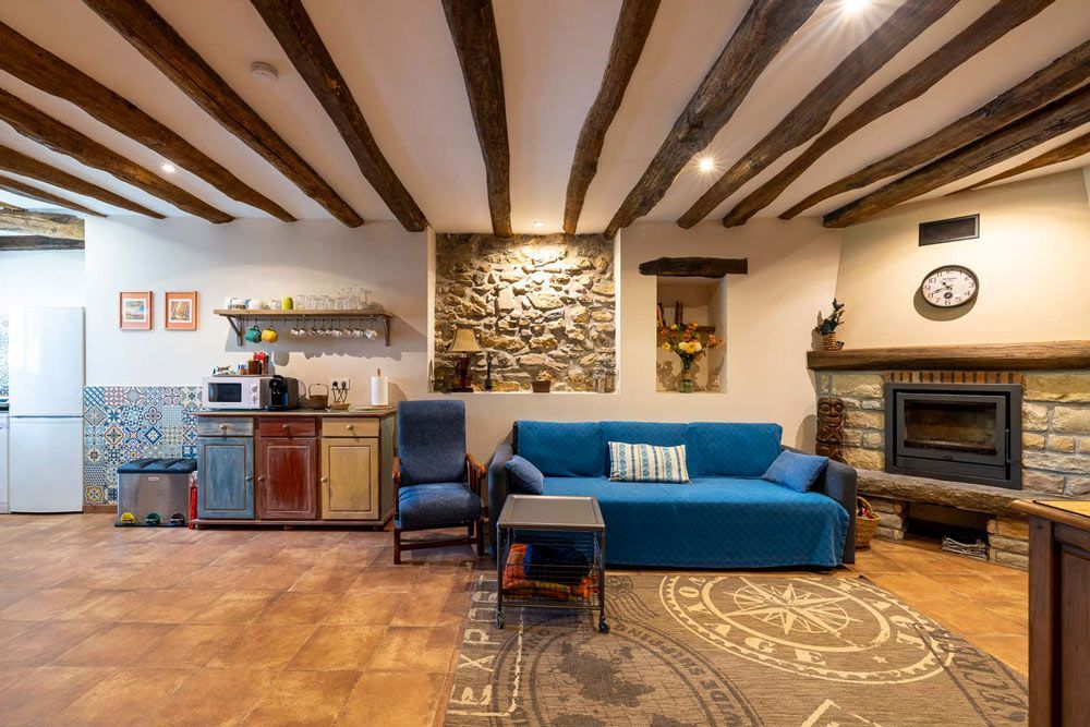 Sala de estar con pared de piedra, vigas a la vista, sofá azul, chimenea y gabinetes rústicos.
