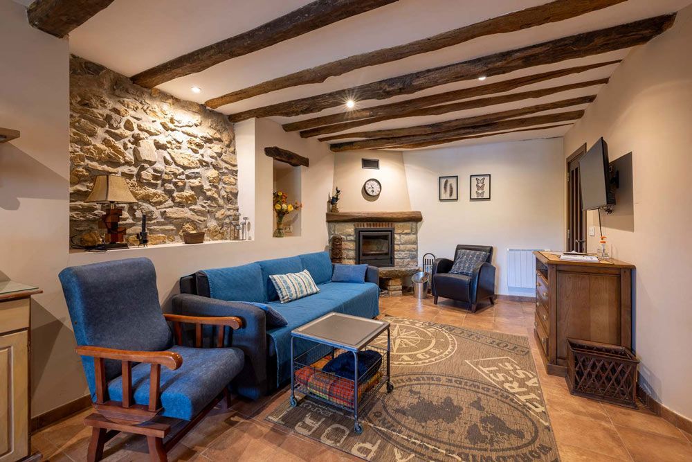 Acogedora sala de estar con pared de piedra, chimenea, sofá azul y vigas de madera a la vista.