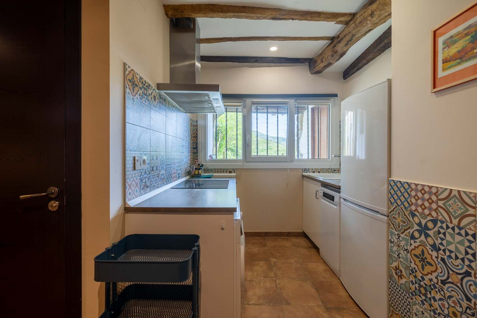 Cocina pequeña con gabinetes blancos, salpicadero de azulejos estampados, ventana y vigas de madera.