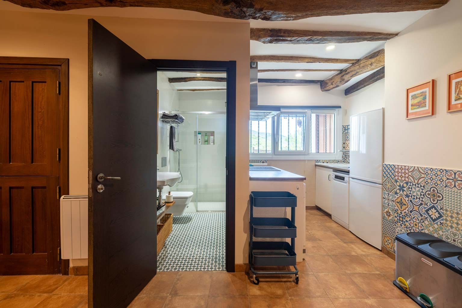 Vista interior: Puerta abierta al baño, cocina con electrodomésticos blancos, vigas rústicas, piso de baldosas.