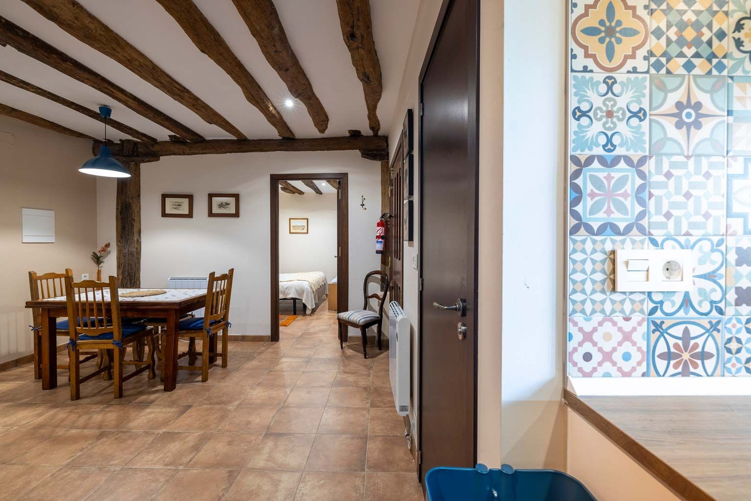 Comedor con vigas de madera, mesa, sillas y pared de azulejos. Se ve la puerta del dormitorio.