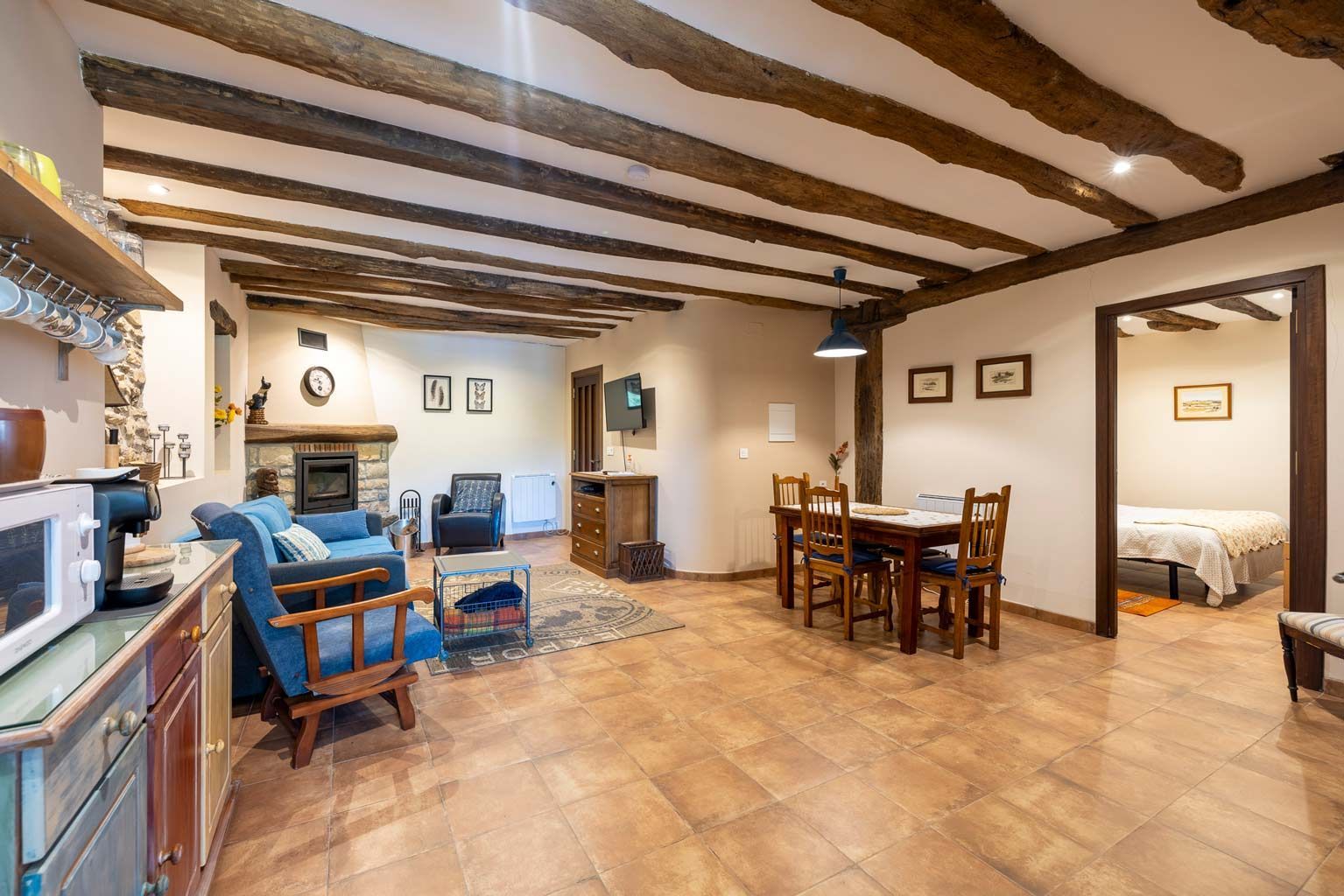 Sala de estar con vigas de madera a la vista, chimenea, comedor y puerta de entrada al dormitorio.