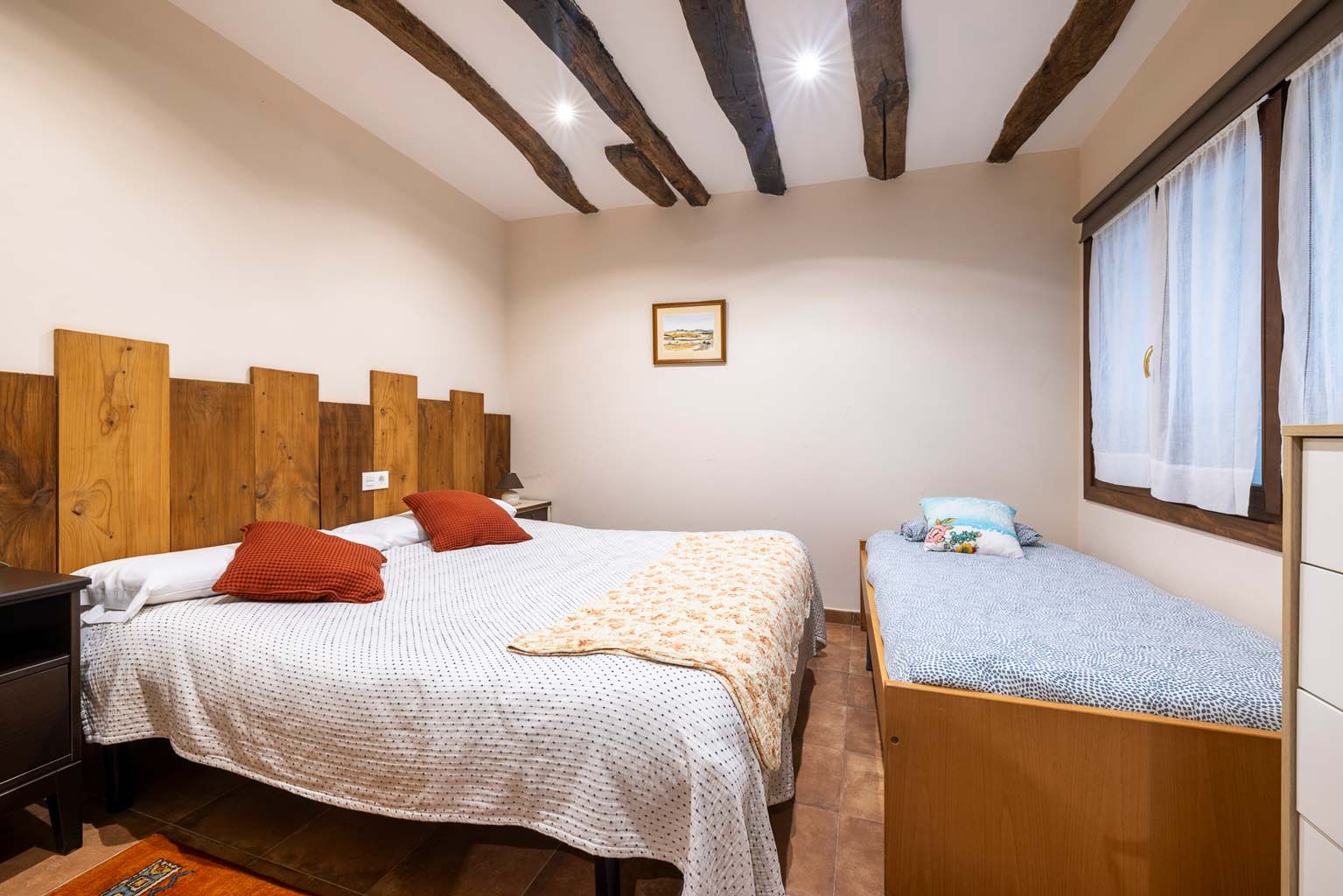 Dormitorio con una cama doble y una cama individual; vigas de madera a la vista en el techo, paredes neutras y una ventana.
