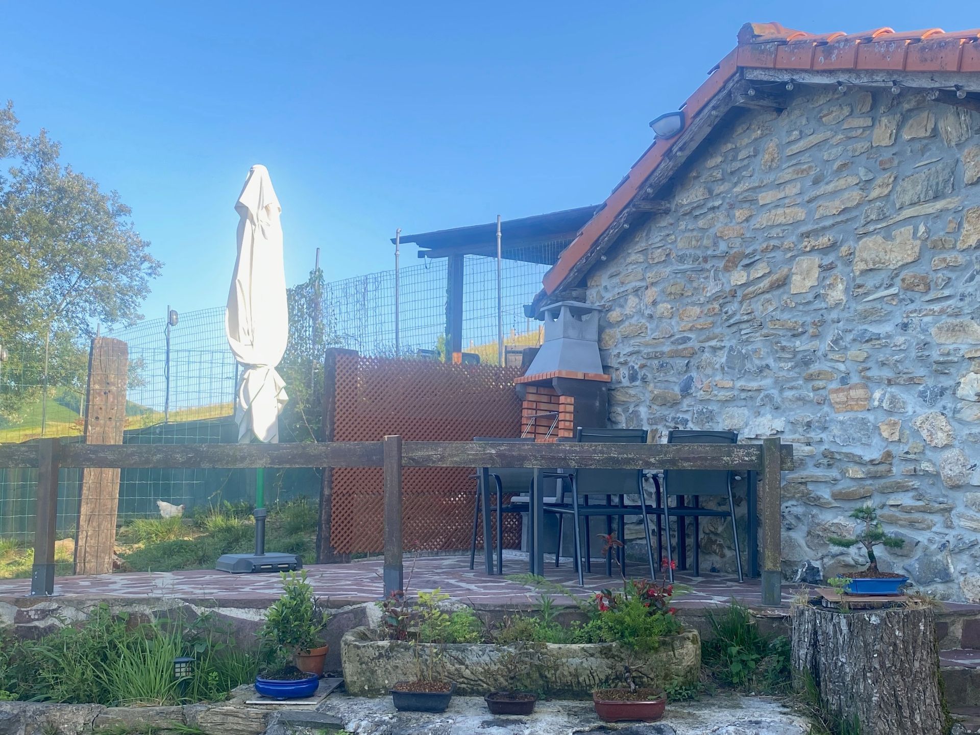Exterior de casa de piedra con patio, parrilla, mesa, sillas y sombrilla bajo un cielo azul.