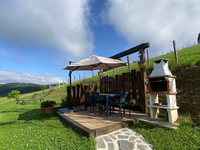 Comedor al aire libre con parrilla, mesa, sombrilla, cerca de madera y colinas verdes bajo un cielo azul con nubes.