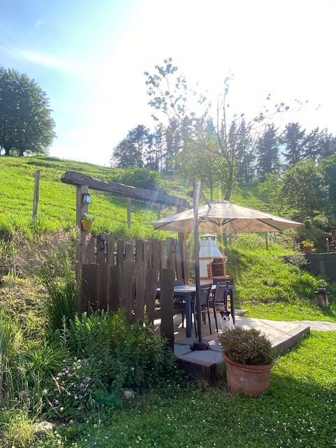 Comedor al aire libre con valla de madera, mesa y sombrilla, en una ladera con césped. Luz solar brillante.