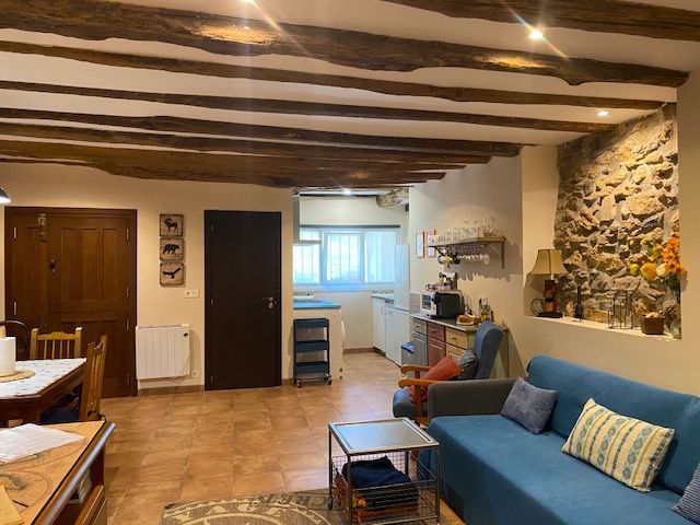 Acogedor espacio de estar con vigas de madera a la vista, pared de piedra, sofá azul y cocina abierta.