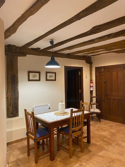 Comedor con mesa y sillas de madera, vigas a la vista, lámpara colgante azul y suelo de baldosas.