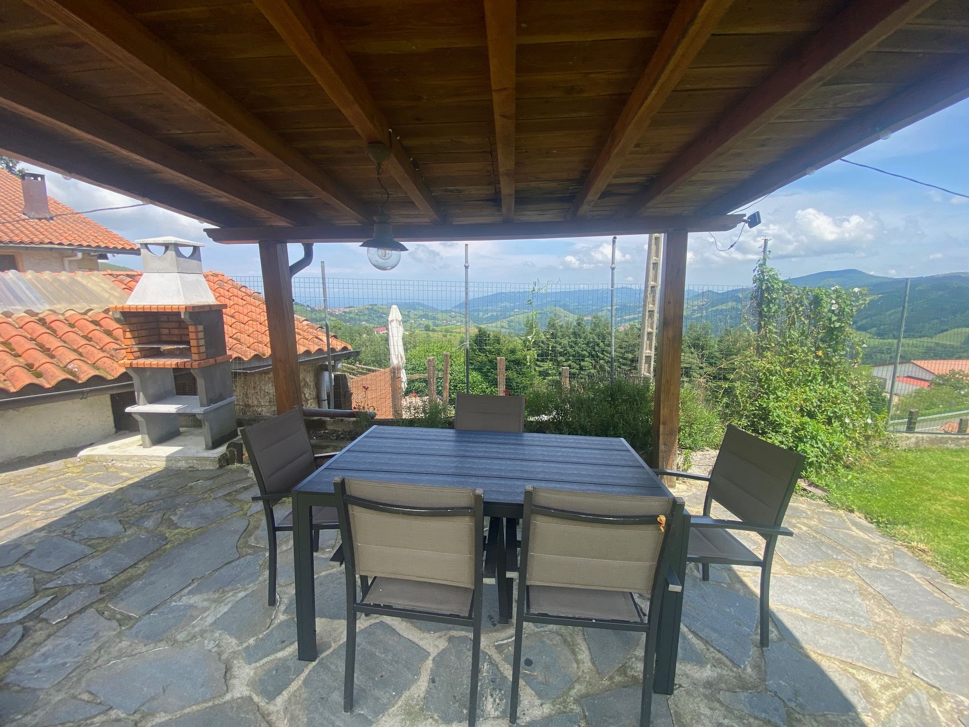 Patio con mesa y sillas bajo techo de madera, con vista a un paisaje montañoso.