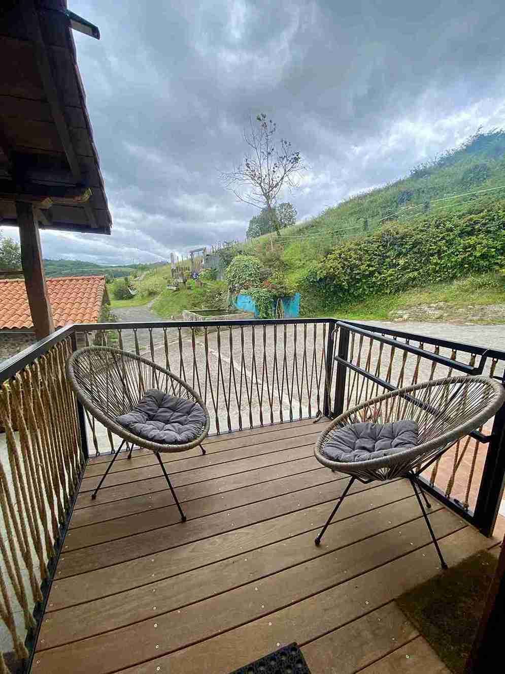 Dos sillas de mimbre en una terraza de madera con vistas a una ladera cubierta de hierba, bajo un cielo nublado.