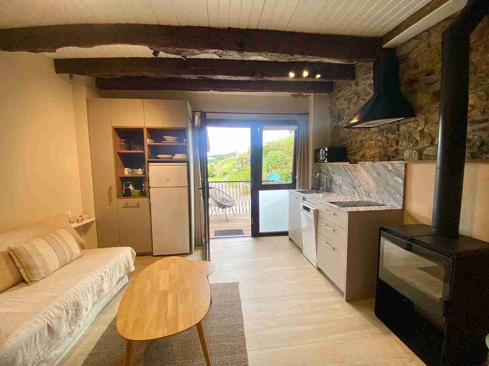 Acogedora cocina con pared de piedra, abierta al patio. Sofá, estufa de leña y armarios en un espacio reducido.