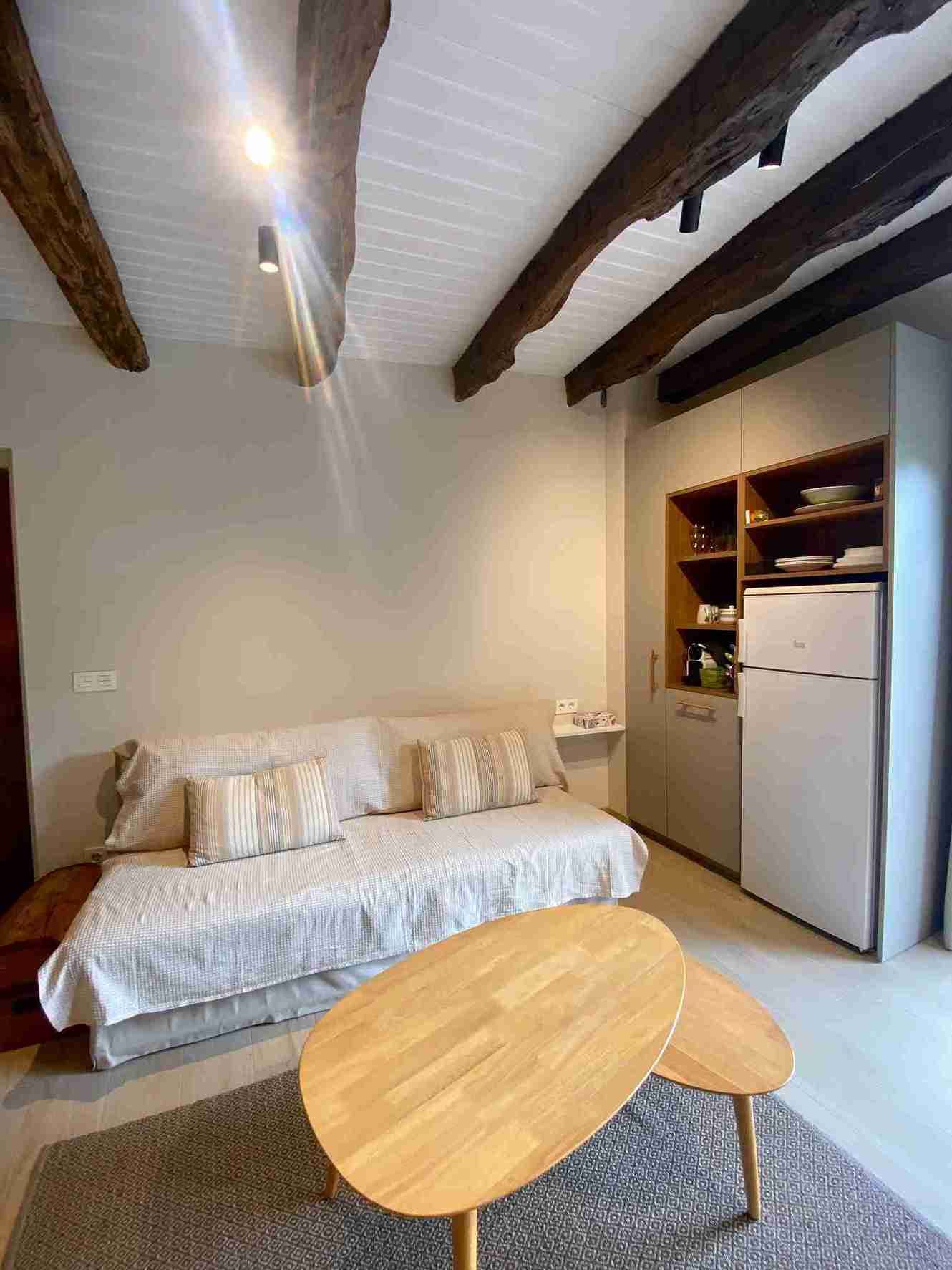 Acogedora sala de estar con sofá, mesa de centro de madera y vigas vistas. Cocina americana con nevera y estanterías.