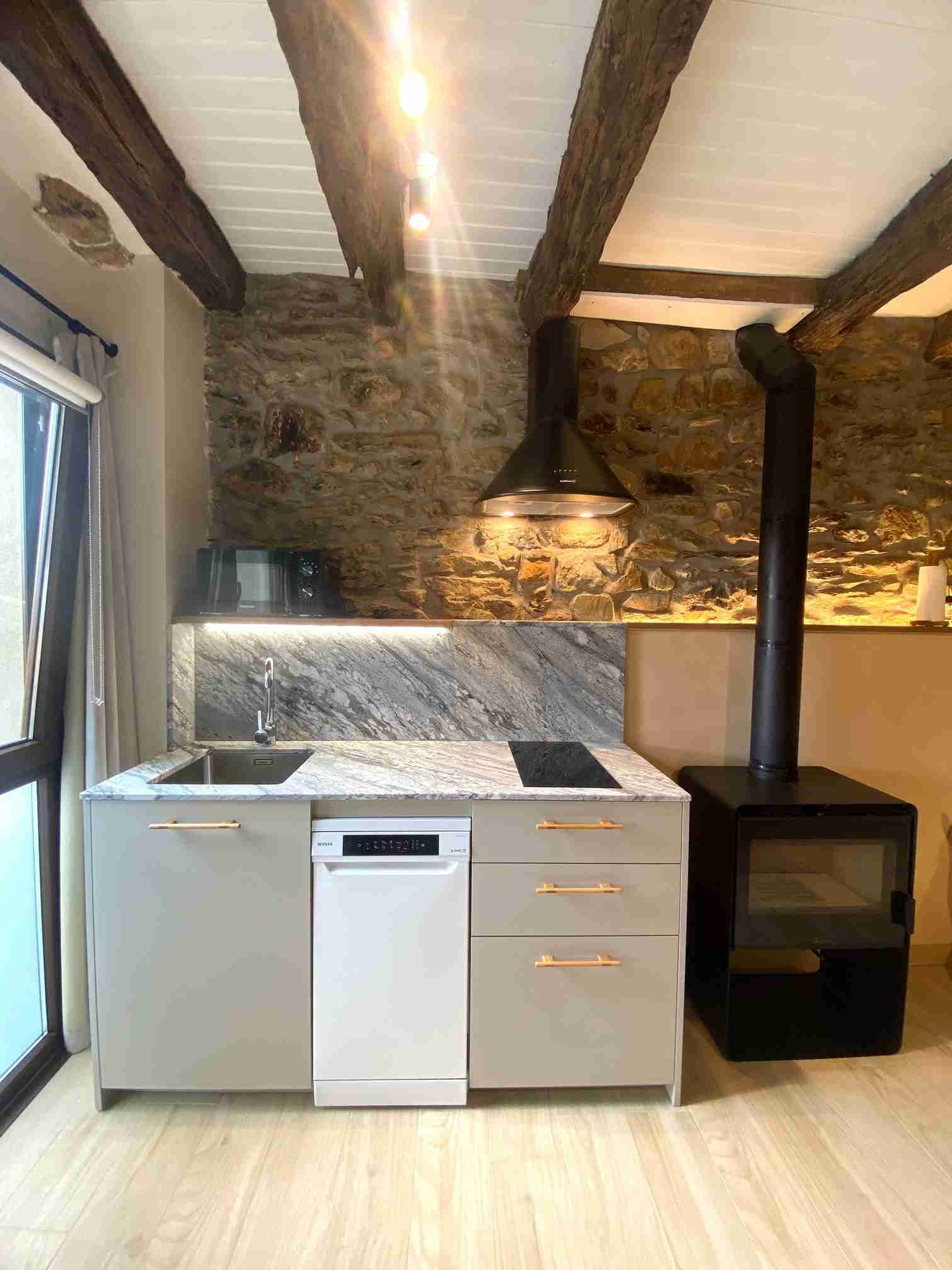 Acogedora cocina con pared de piedra, vigas de madera y una pequeña estufa de leña. Gabinetes gris claro y encimera de granito.
