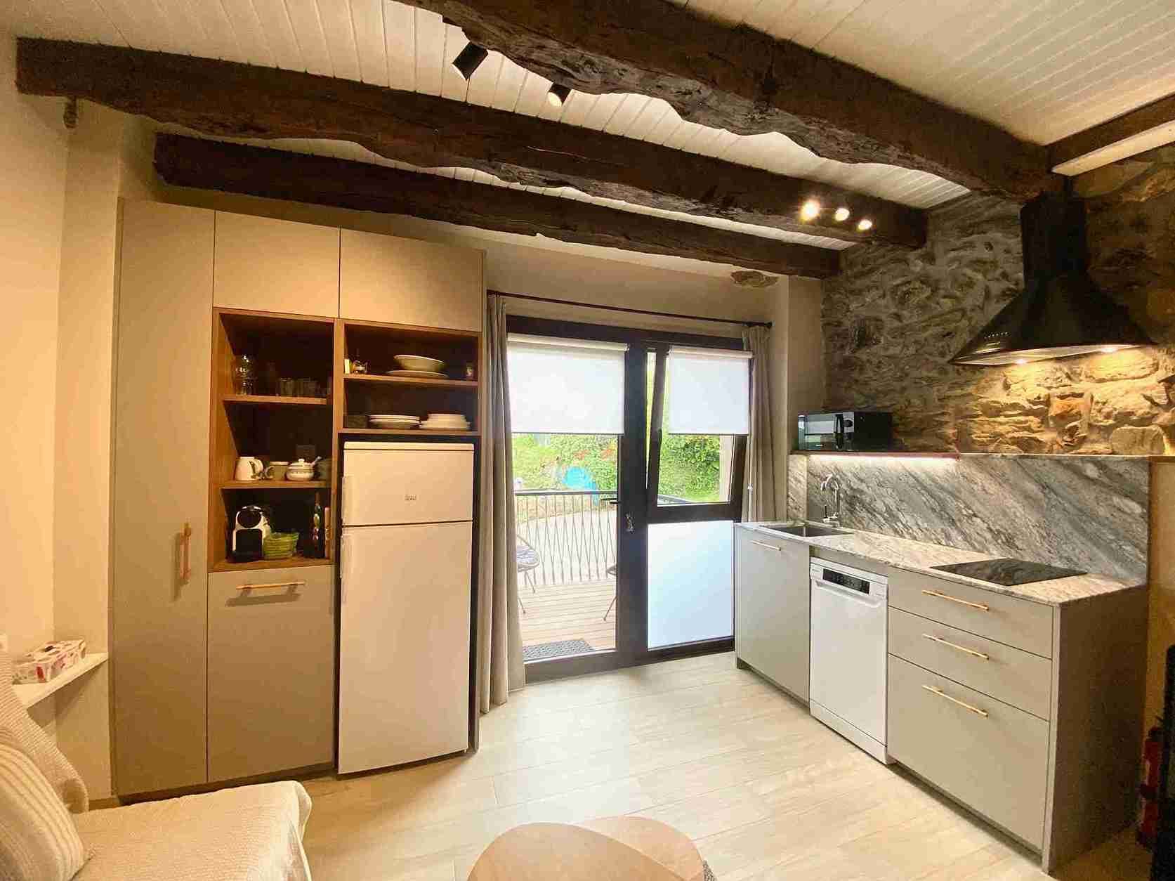Acogedora cocina con pared de piedra, vigas de madera y electrodomésticos modernos. Una ventana da a una pequeña zona exterior.