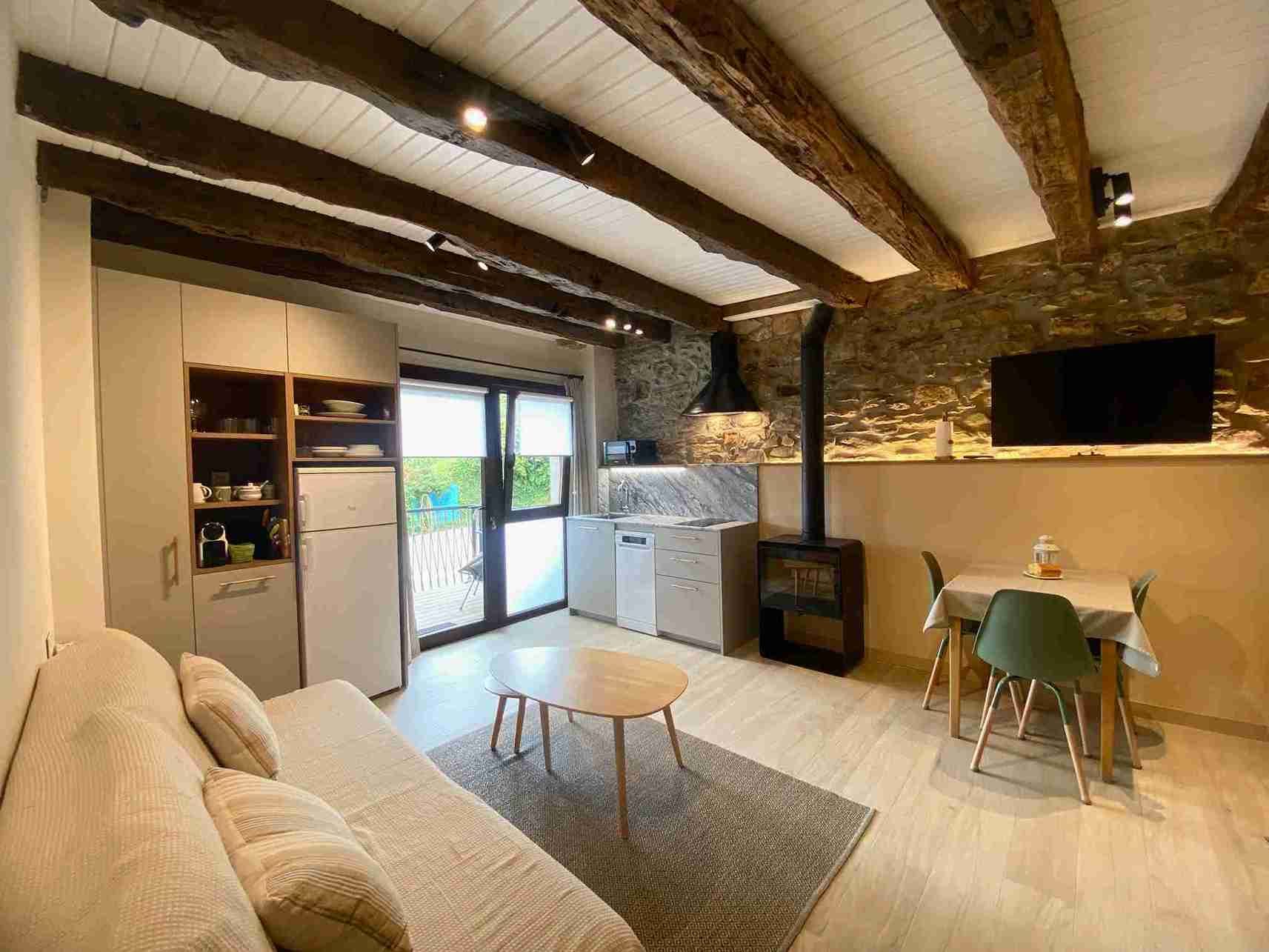 Acogedor apartamento tipo estudio con vigas a la vista, pared de piedra, cocina americana, sofá y mesa de comedor.