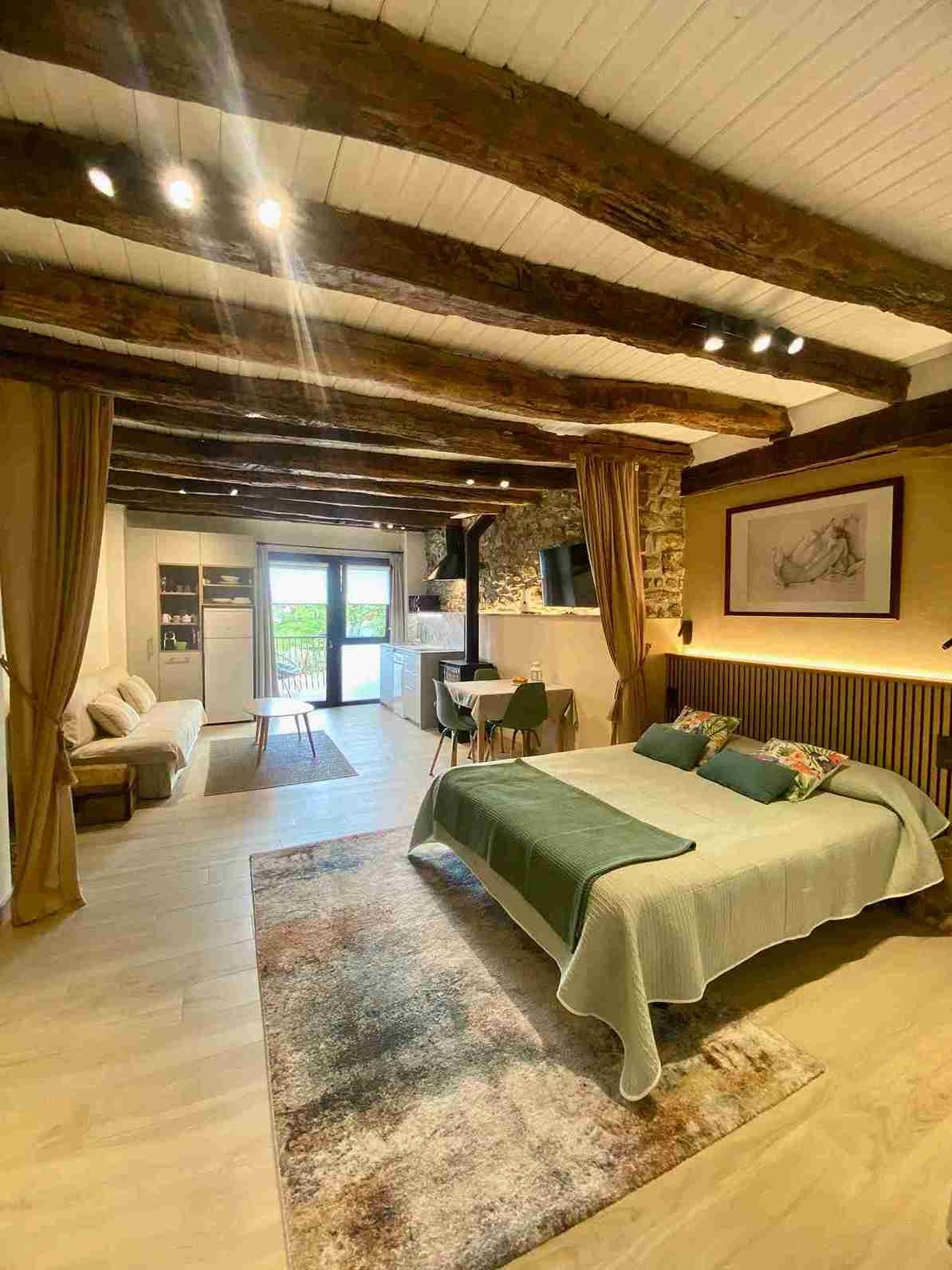 Acogedor dormitorio con vigas vistas, cama, sofá y chimenea. Tonos tierra y luz natural.