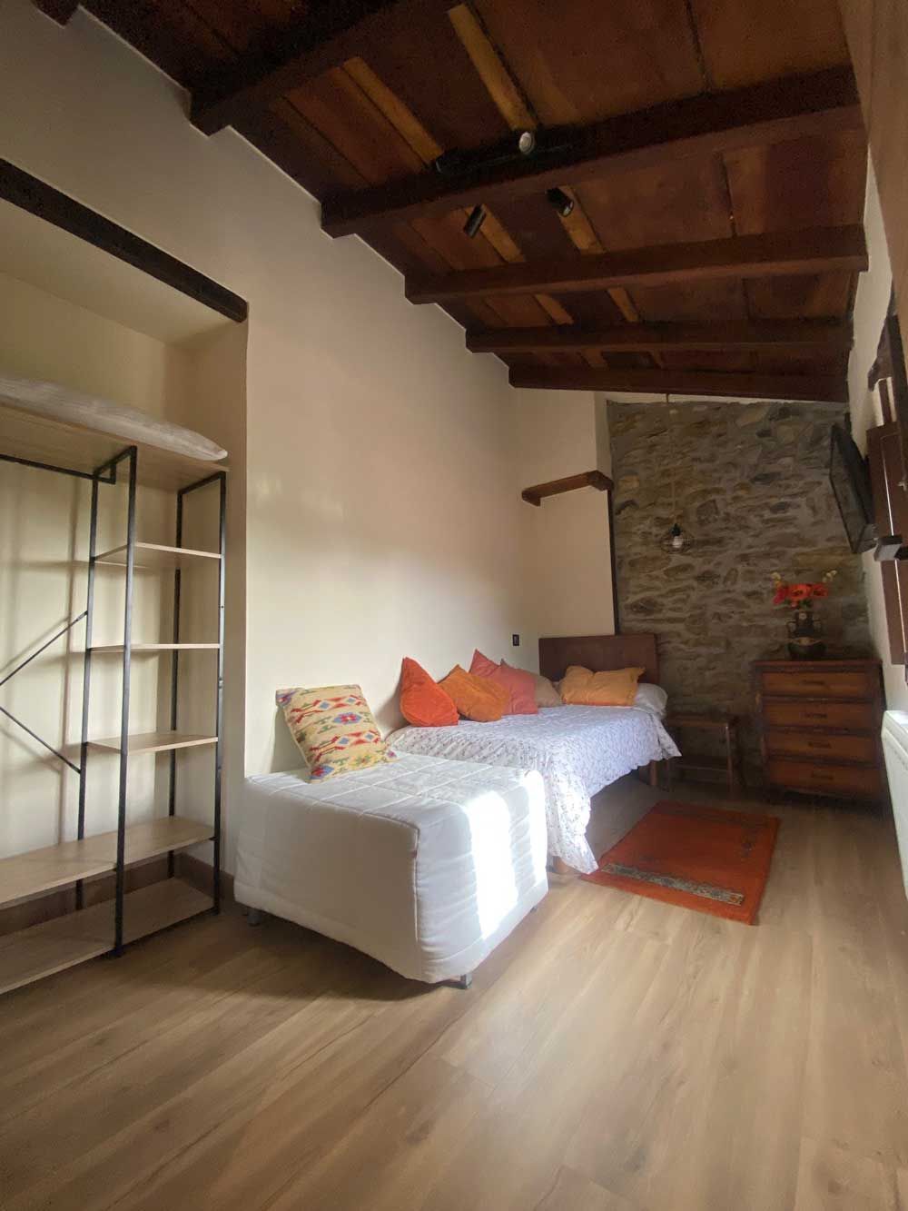 Pequeño dormitorio con cama, techo de madera, pared de piedra y estantería abierta.
