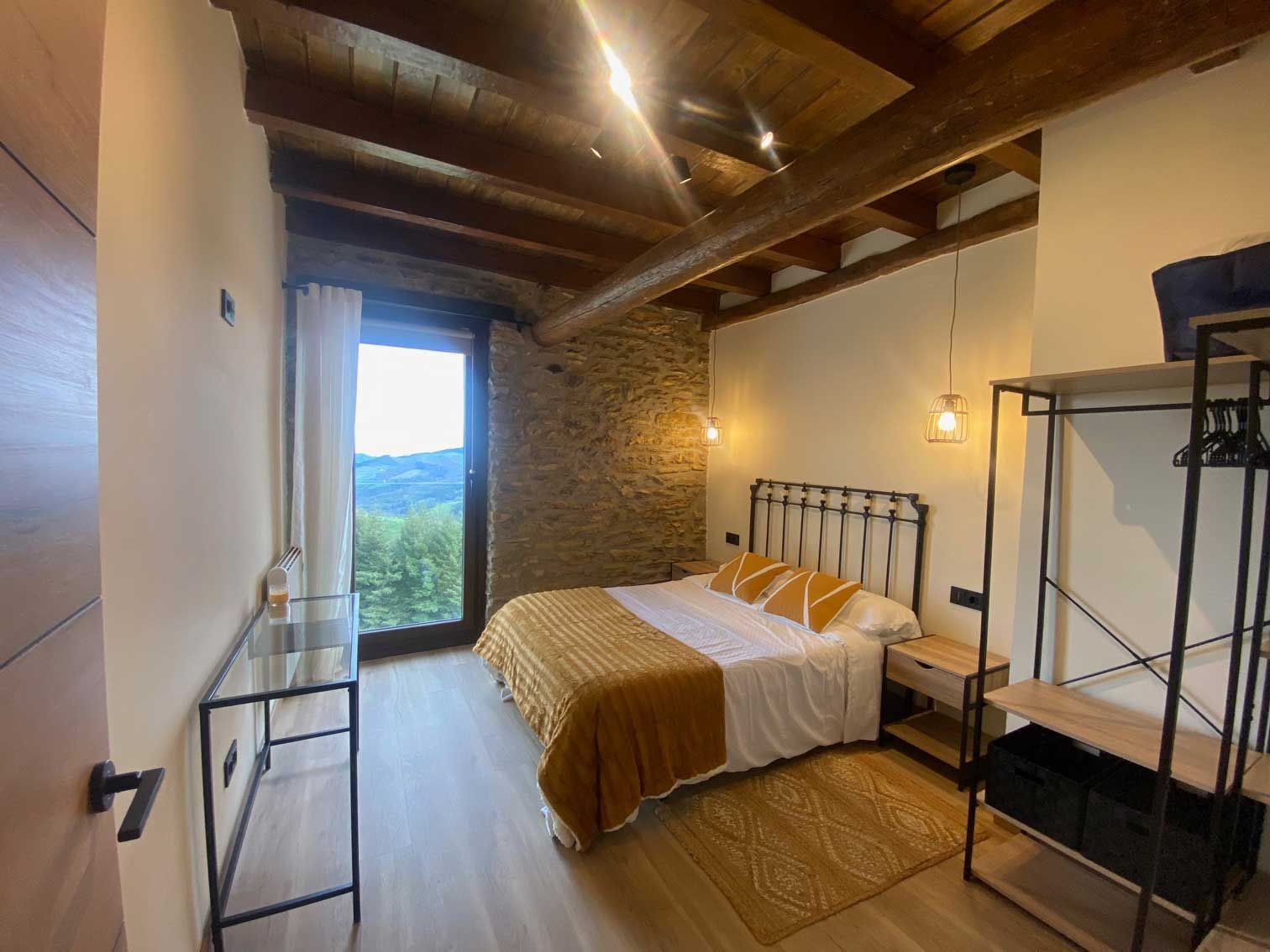 Dormitorio con vigas a la vista, pared de piedra, cama con detalles en amarillo, armario abierto y ventana con vista panorámica.
