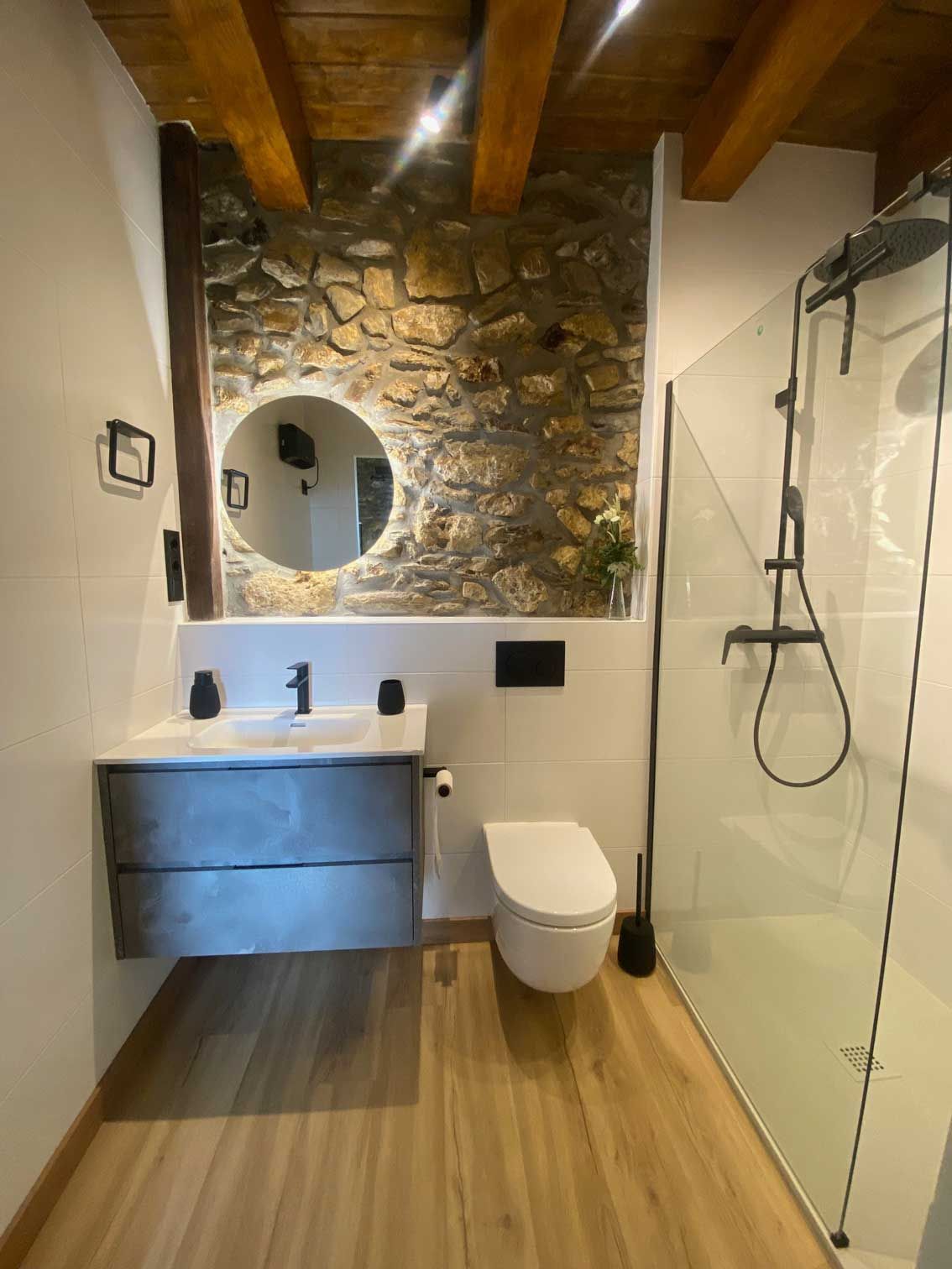 Baño con pared de piedra, vigas de madera, tocador moderno, inodoro y ducha de cristal.