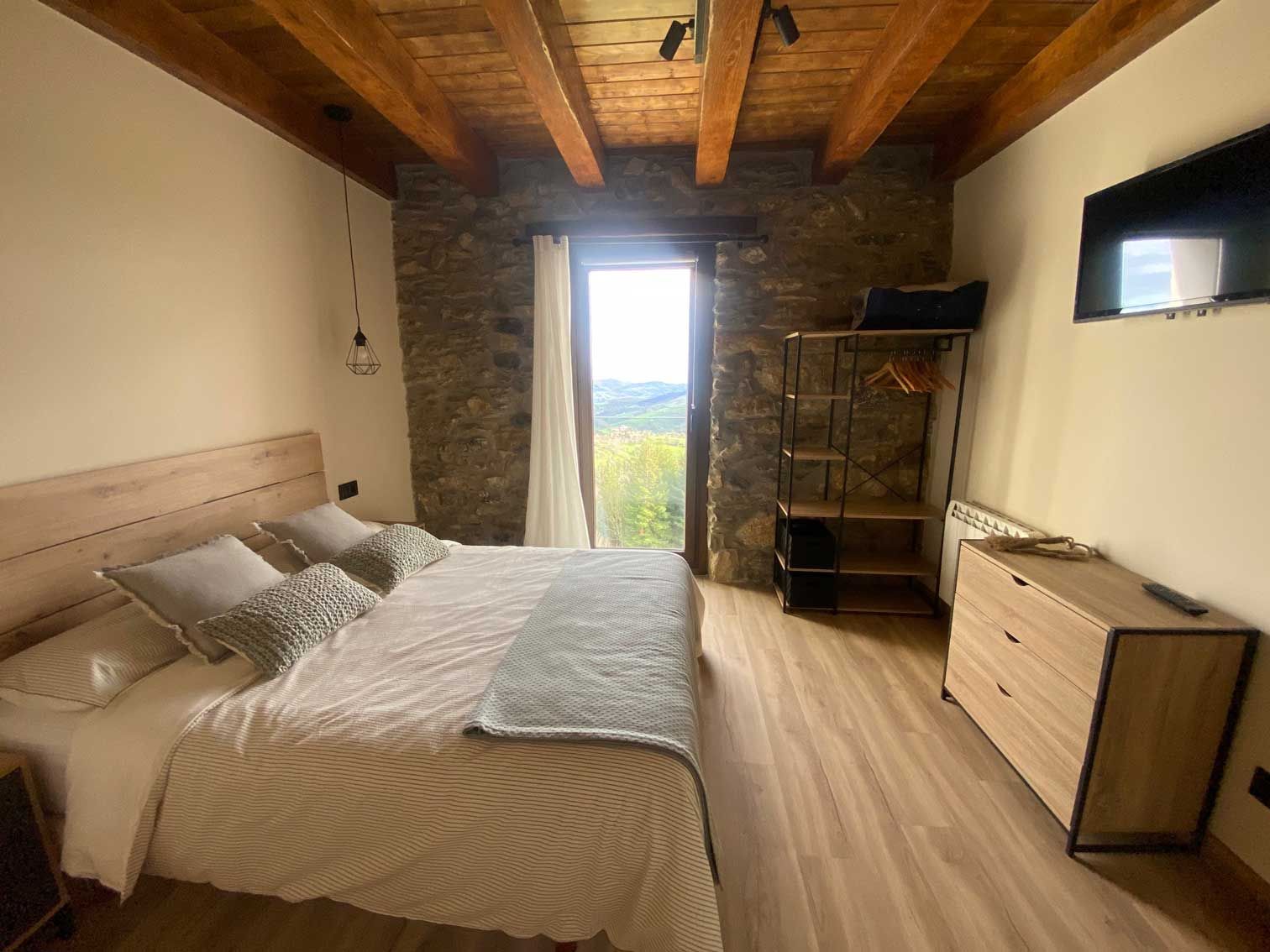 Dormitorio con cama, pared de piedra, ventana con vista, cómoda y TV.