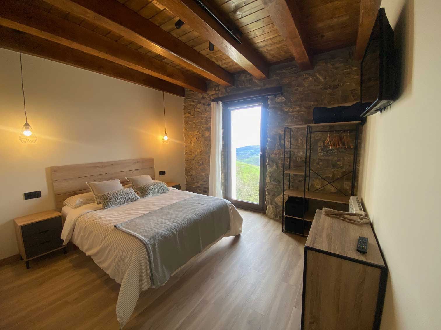Un dormitorio con una cama, mesita de noche, televisión y un gran ventanal.