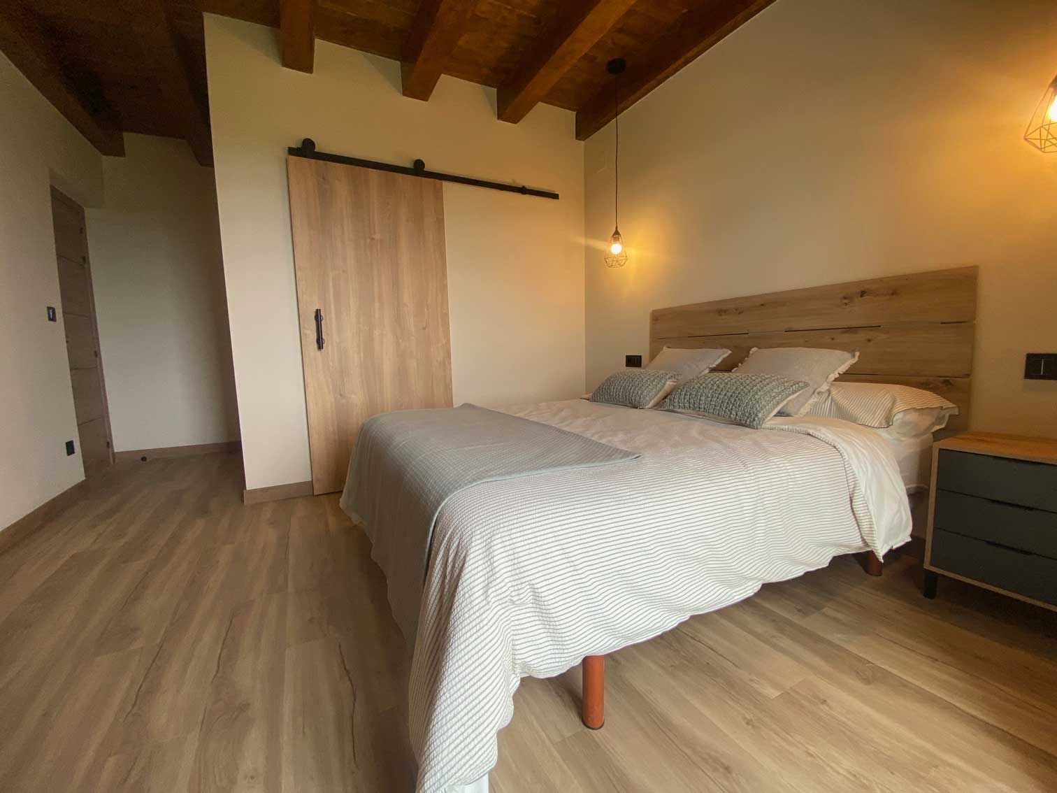 Dormitorio con cama, puerta corrediza, suelo de madera y techo con vigas a la vista.