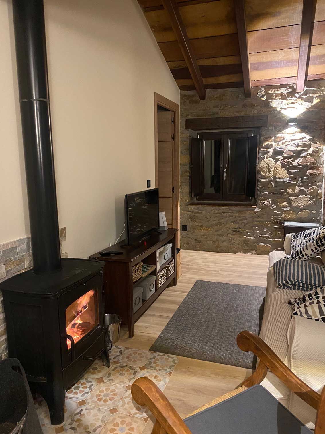 Una acogedora sala de estar con chimenea encendida, televisor sobre un soporte, pared de piedra y una ventana.