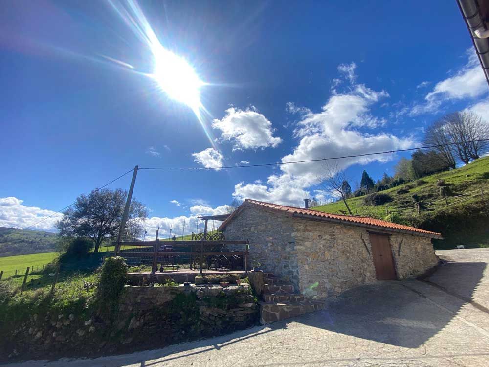 Cabaña de piedra en un día soleado con cielo azul, nubes blancas y ladera verde.