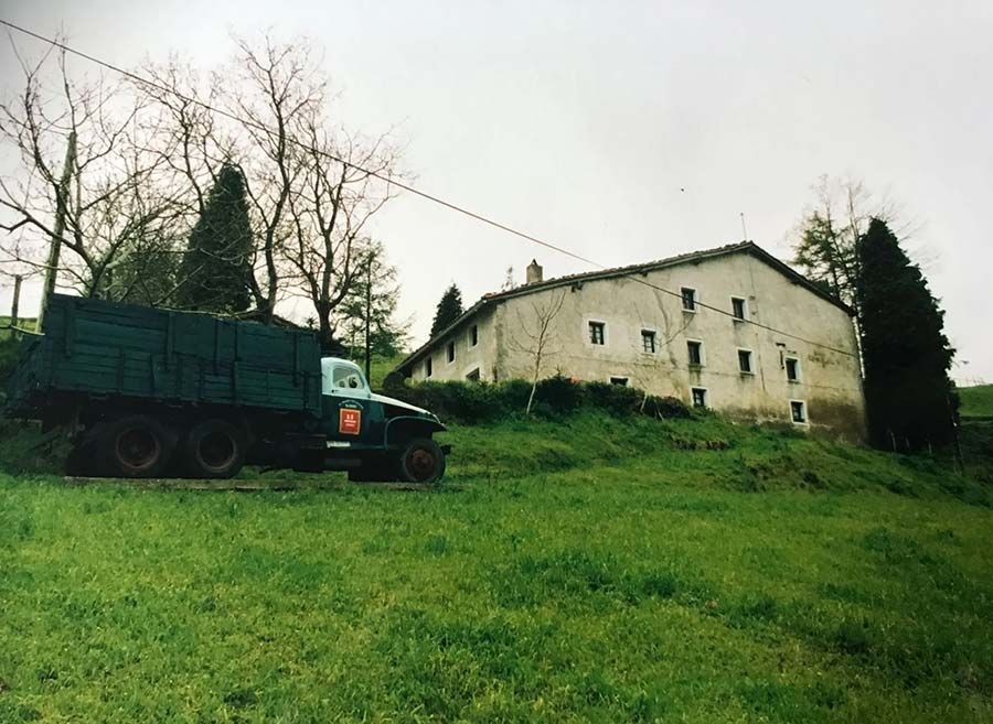 Camión verde estacionado junto a una casa de campo blanca en una colina cubierta de hierba bajo un cielo nublado.