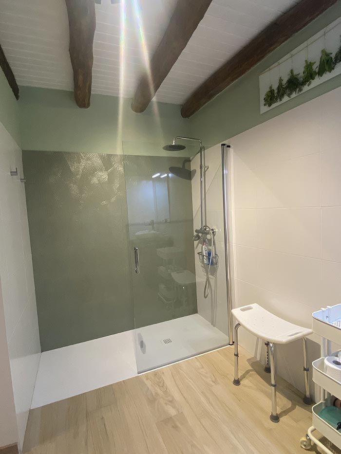 Ducha en un baño con pared verde, vigas de madera y silla de ducha plegable a la derecha.