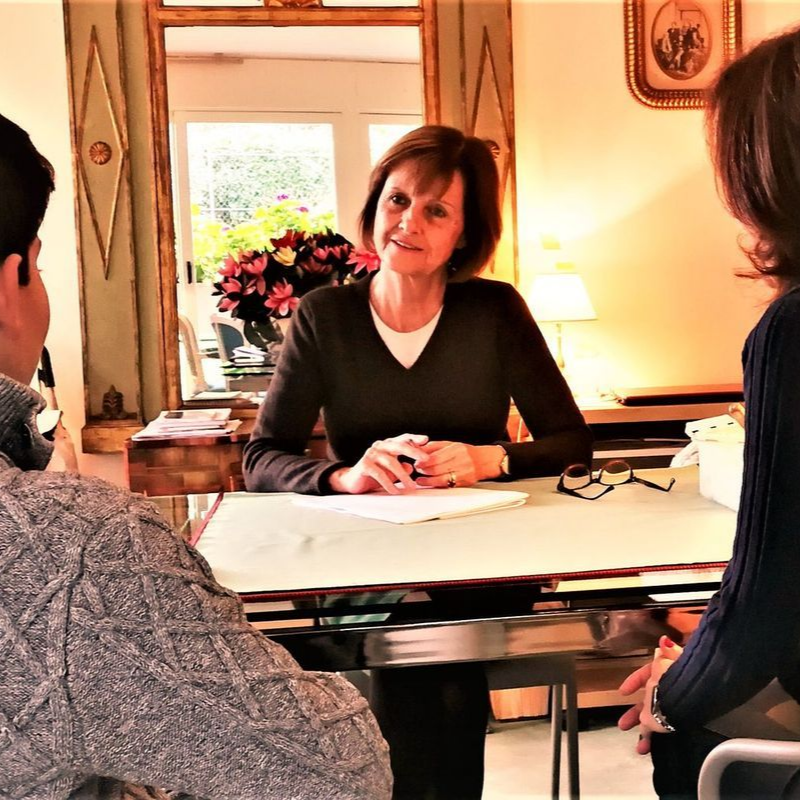 Une femme est assise à une table avec deux personnes, des fleurs et une fenêtre sont visibles.