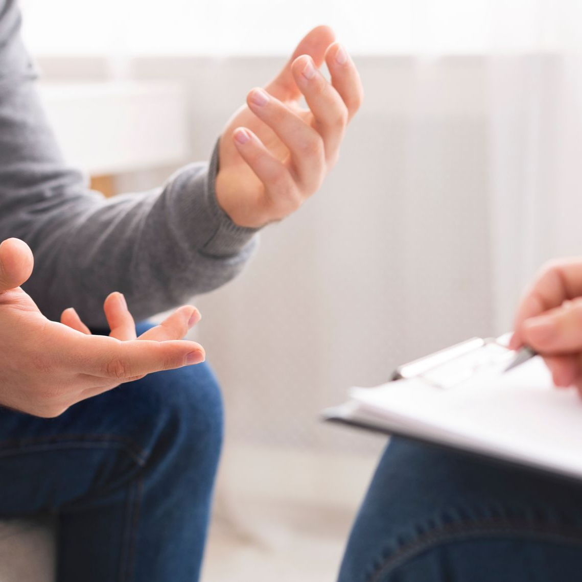 Une personne gesticule en parlant à quelqu'un qui tient un bloc-notes. Dans une pièce.