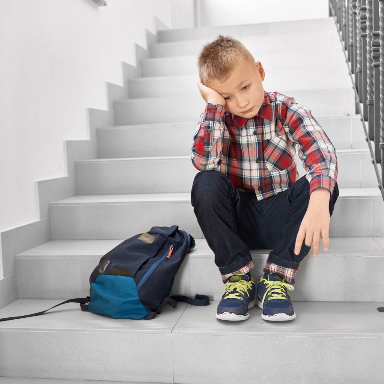 Un garçon est assis l'air abattu sur des marches, avec un sac à dos ; chemise à carreaux, jean foncé, baskets bleues.