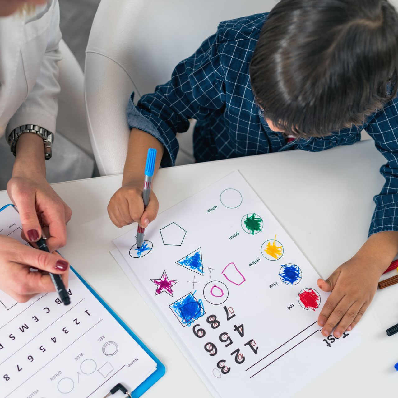 Une personne et un enfant travaillent sur une fiche d'activités. L'enfant colorie des formes et des chiffres avec un stylo.