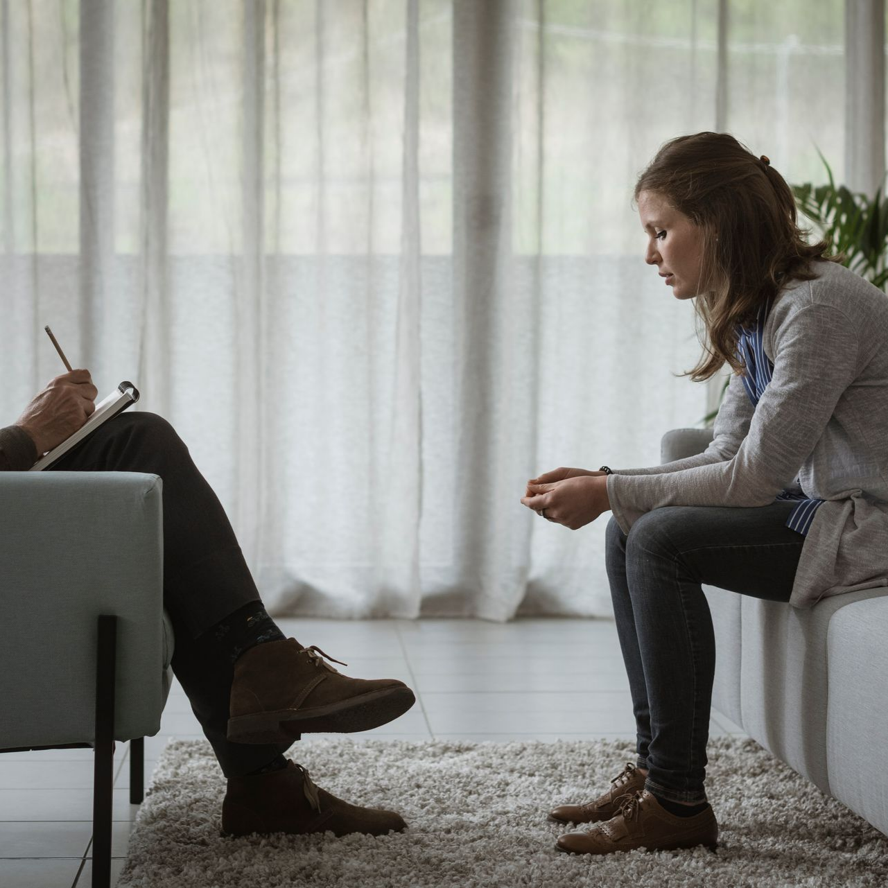 Une femme en thérapie, assise sur un canapé, parle à une personne qui écrit sur un bloc-notes dans une pièce avec une fenêtre.