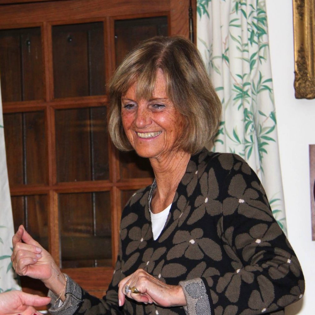 Femme souriante, la main levée. Elle porte un pull foncé à motifs floraux. Elle se tient près d'une fenêtre en bois et d'un rideau.