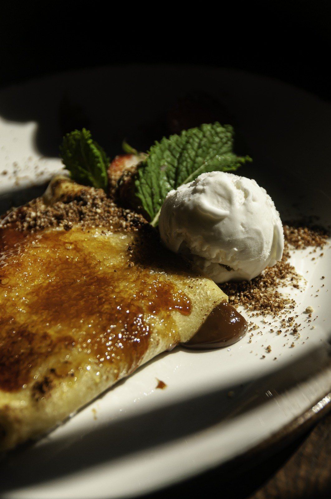 Crepe con azúcar caramelizado, cubierto con migas de chocolate, helado, una rodaja de fresa y menta en un plato blanco.