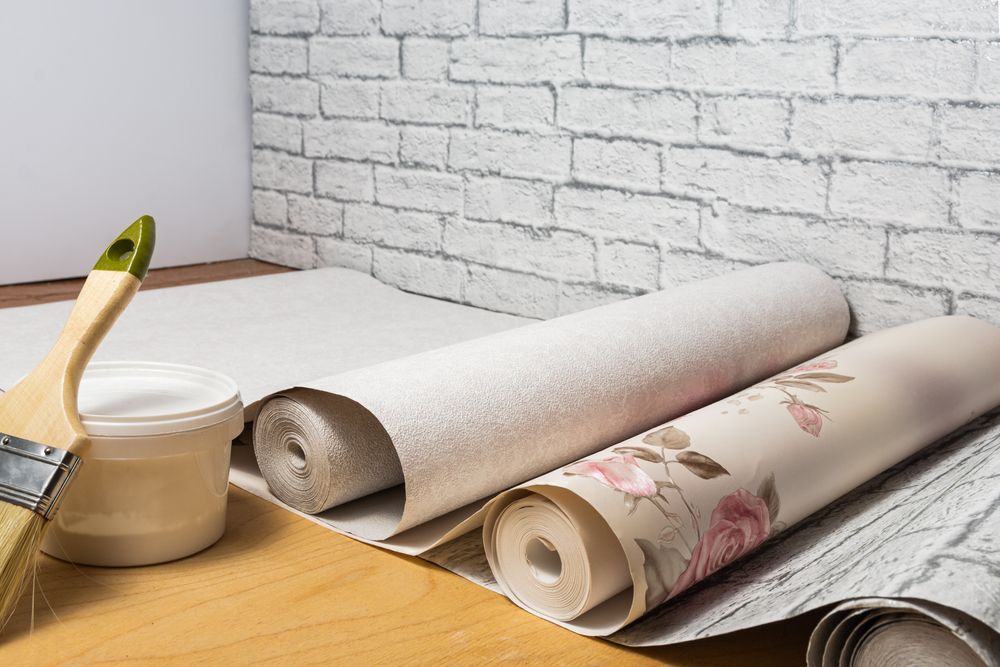 Rollos de papel tapiz, pincel y adhesivo sobre una superficie, junto a una pared parcialmente empapelada