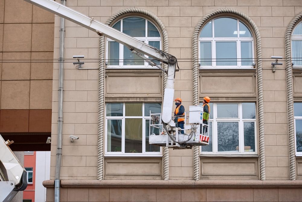 Dos trabajadores en un elevador aéreo reparan la fachada de un edificio con ventanas arqueadas.