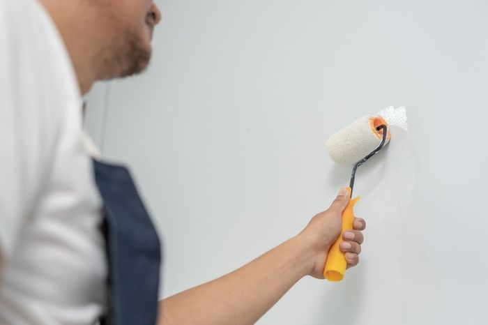 Persona pintando una pared blanca con un rodillo, vestida con camisa blanca y delantal azul.