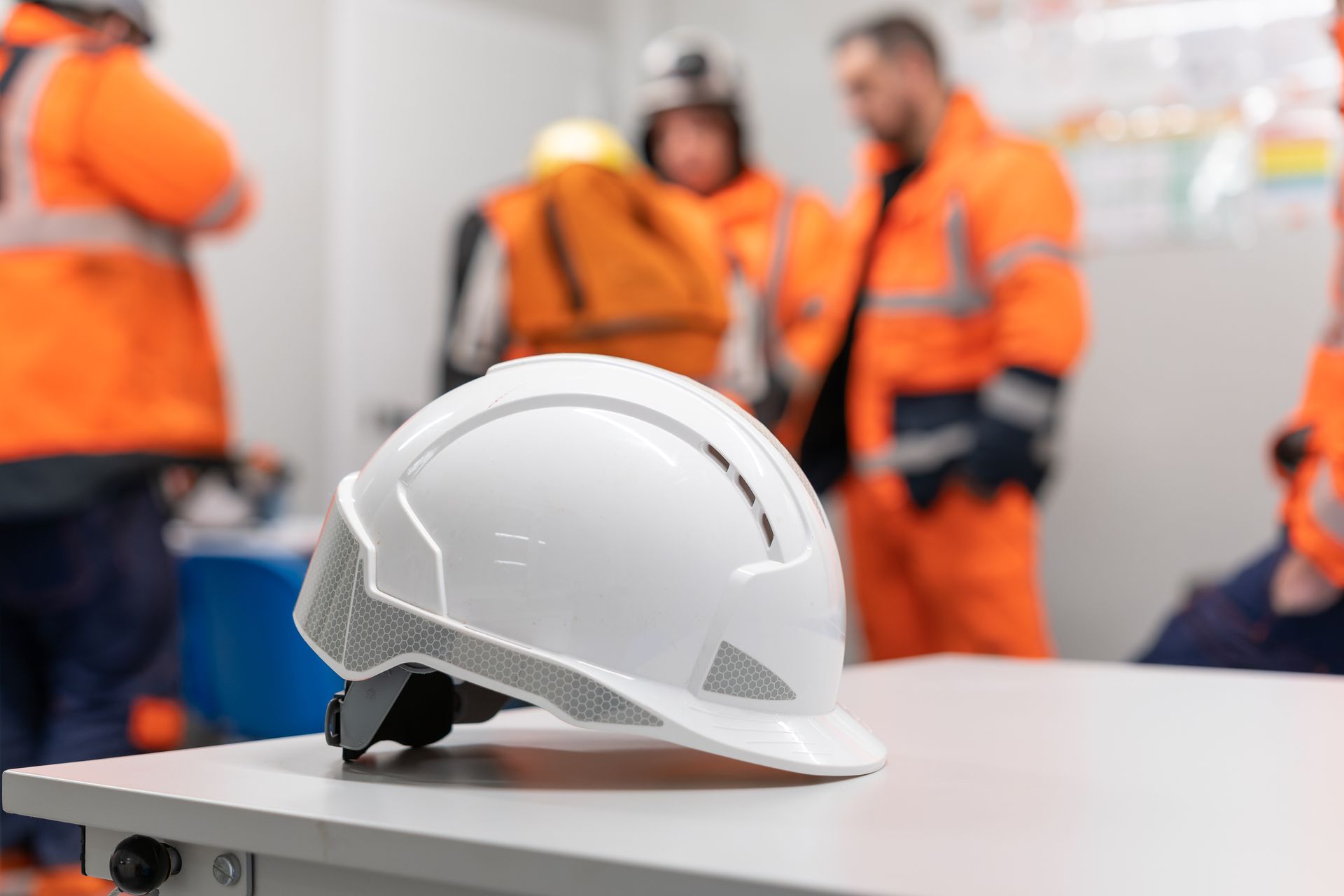 Casque de chantier blanc posé sur une table devant des ouvriers flous portant des gilets de sécurité orange.