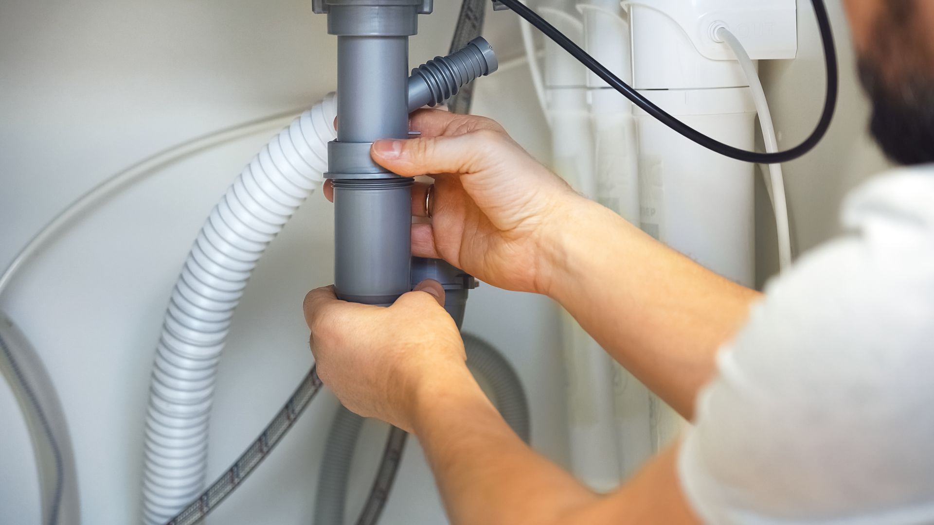 Des mains ajustent un tuyau gris et un raccord de tuyauterie sous un évier ou un appareil électroménager.