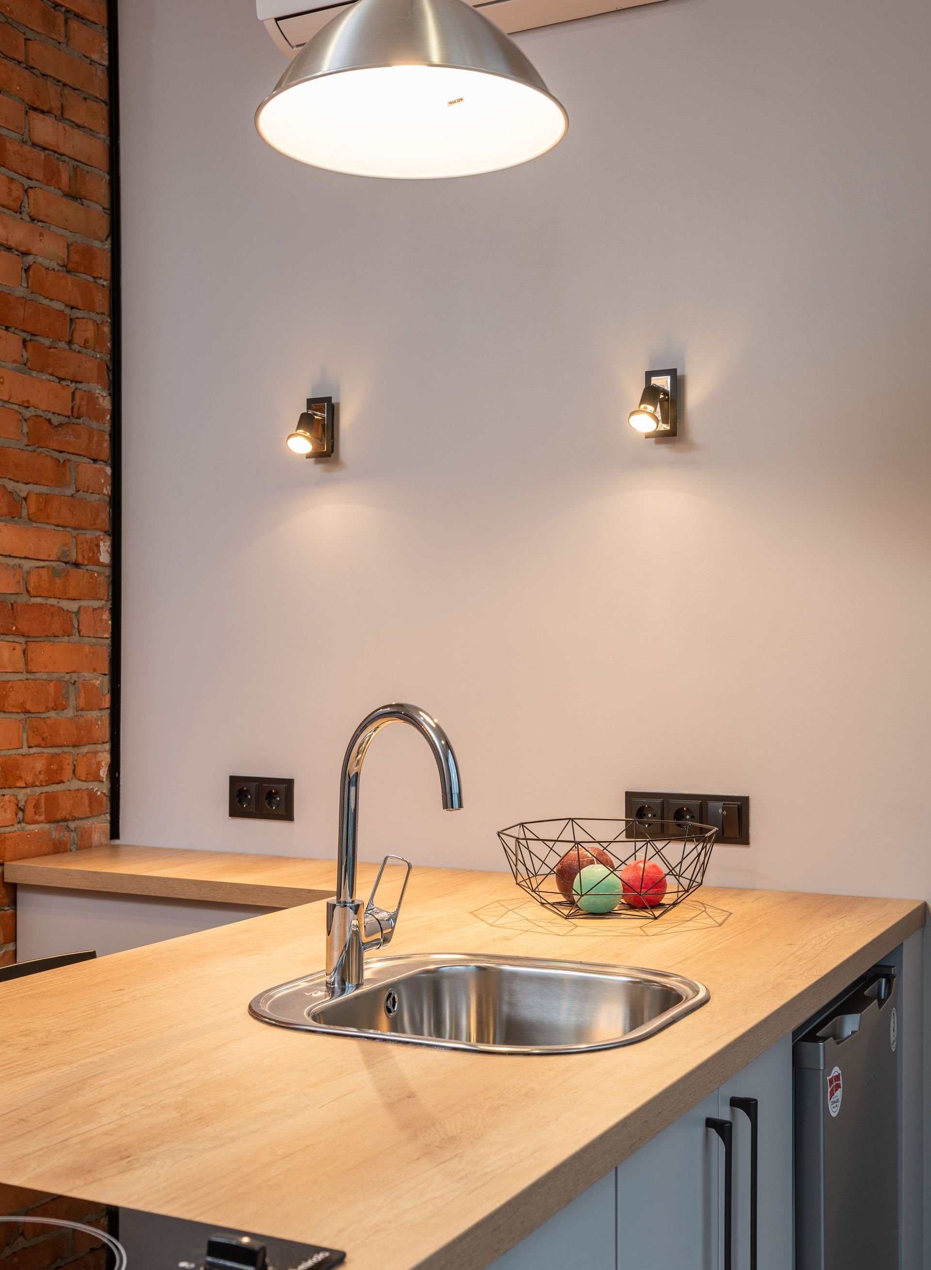 Plan de travail de cuisine moderne avec évier en acier inoxydable, surface en bois, suspension lumineuse et mur en briques apparentes