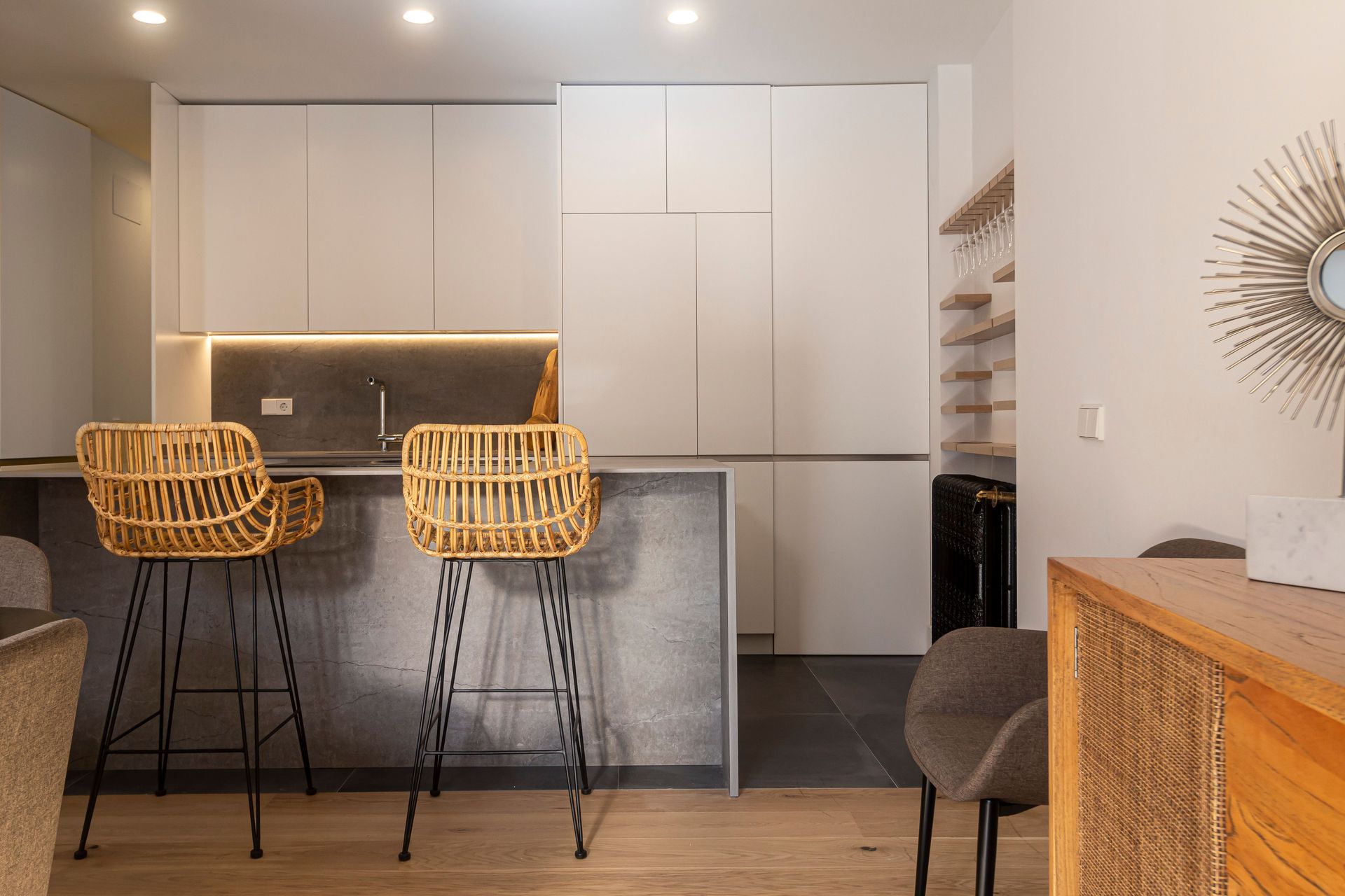 Cuisine moderne avec placards blancs, îlot gris et deux tabourets de bar en osier sous des suspensions lumineuses.